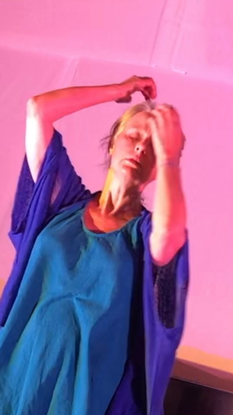 Une personne en robe blanche se produit sur une scène éclairée par un éclairage violet, les mains tendues.