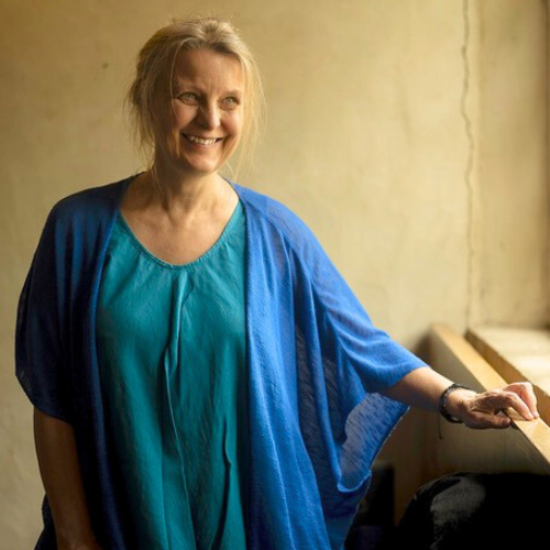 Femme souriante, regardant ailleurs, portant des vêtements bleus, debout près d'une fenêtre.