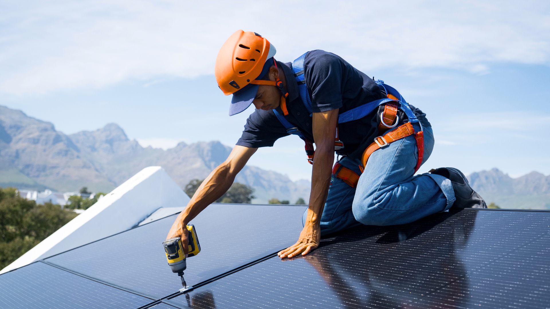 Solar panel installer kneeling on a rooftop, using a power drill. Orange helmet and harness are visible. Mountains in the background.