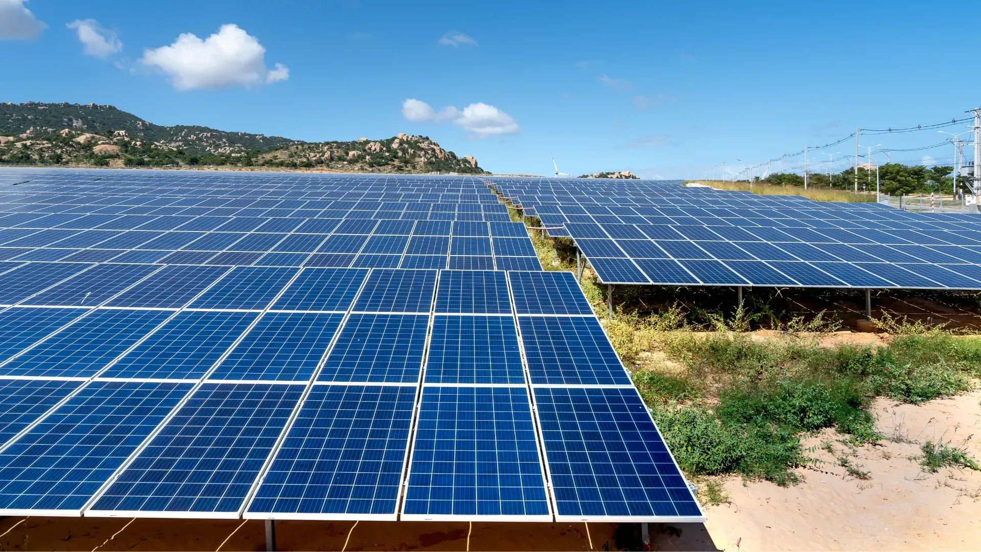 A large field of solar panels stretching under a bright blue sky with scattered clouds and distant hills.