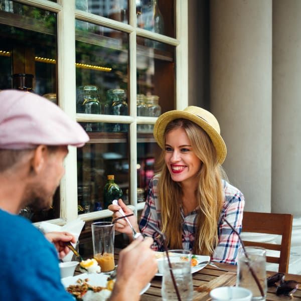 A person in a pink cap and another in a straw hat dine together at an outdoor table with food and drinks.