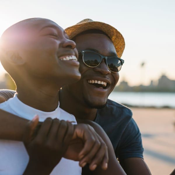 Two smiling people embracing outdoors, bathed in warm sunlight against a blurred waterfront background.