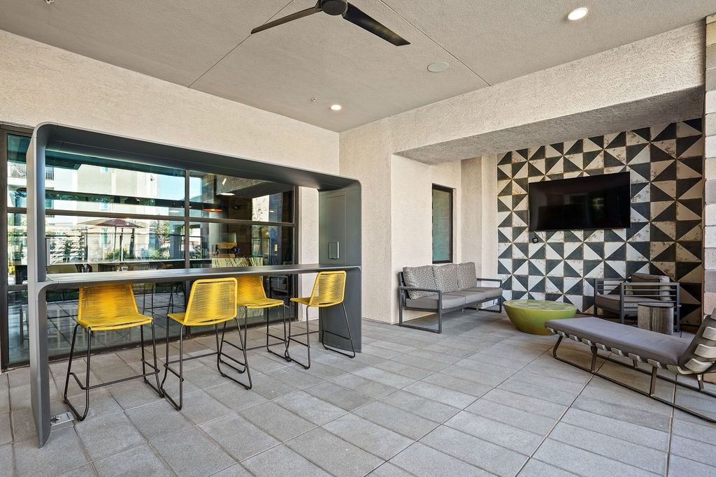Outdoor community lounge with bar-height seating, yellow stools, a sofa, and a wall-mounted TV.