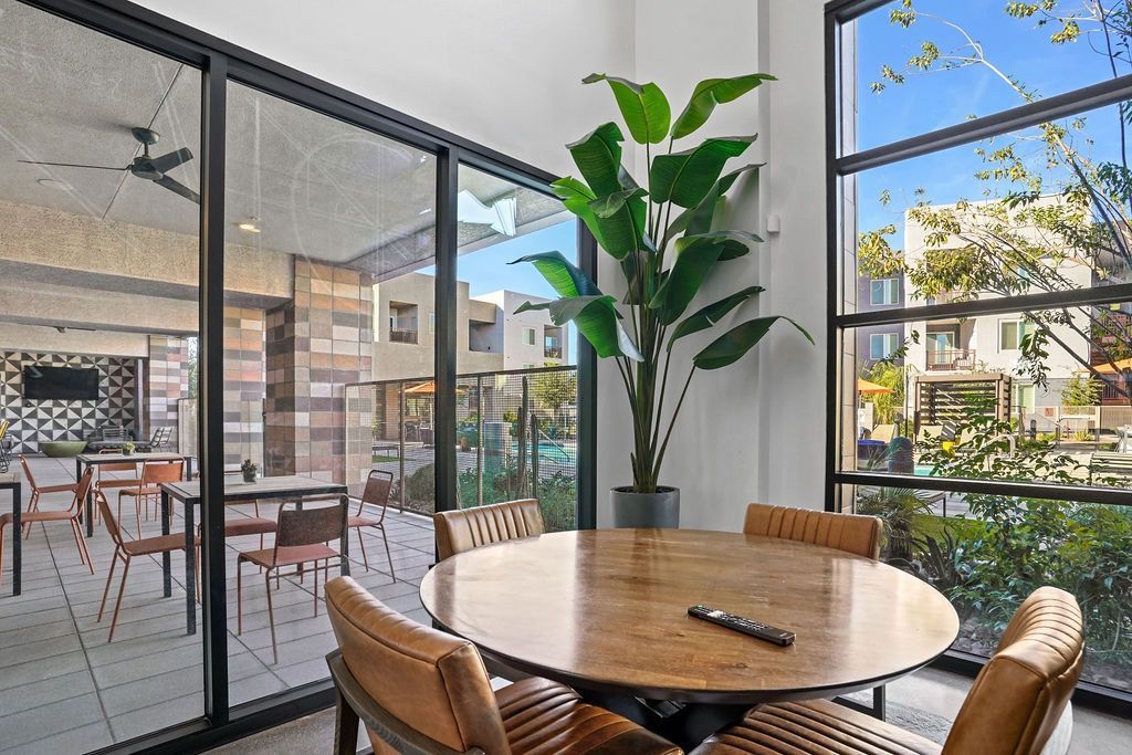 Indoor community lounge with a round table, tan chairs, a tall plant, and glass walls overlooking a pool.