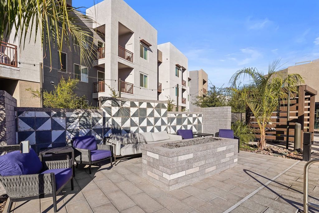 Outdoor communal patio with seating, a central fire pit, and geometric-pattern wall near apartment buildings.
