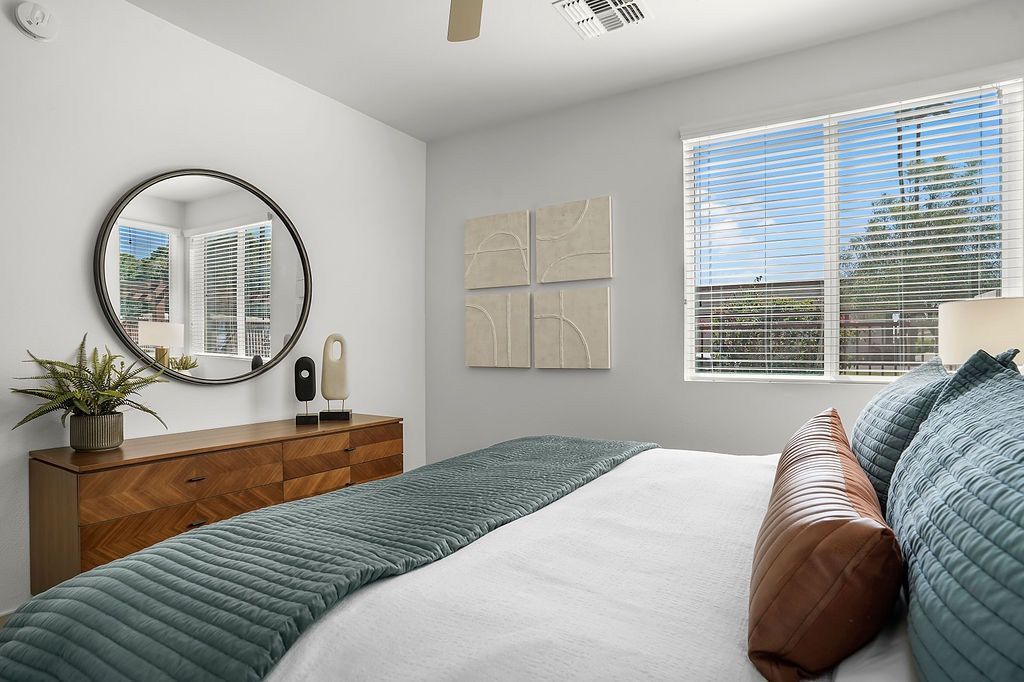 Bedroom with a wooden dresser, round mirror, plants, and a bed by a large window with blinds.