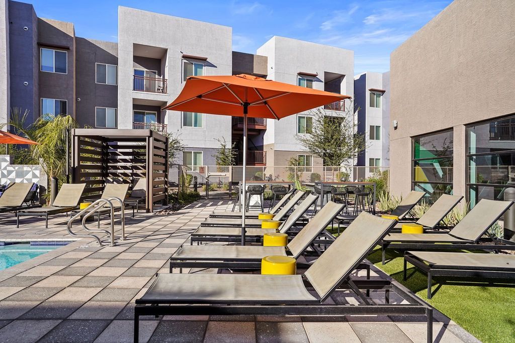 Outdoor pool area with lounge chairs, orange umbrella, and modern apartment buildings.