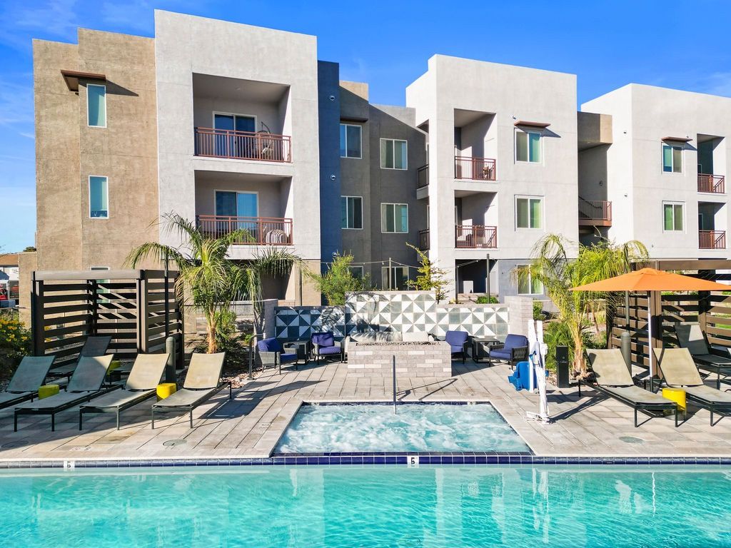 Outdoor pool area with lounge chairs, seating, and palm trees in front of modern apartment buildings.