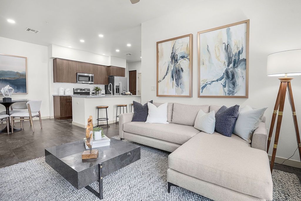 Open-concept living area with beige sectional sofa, coffee table, and kitchen visible in background.