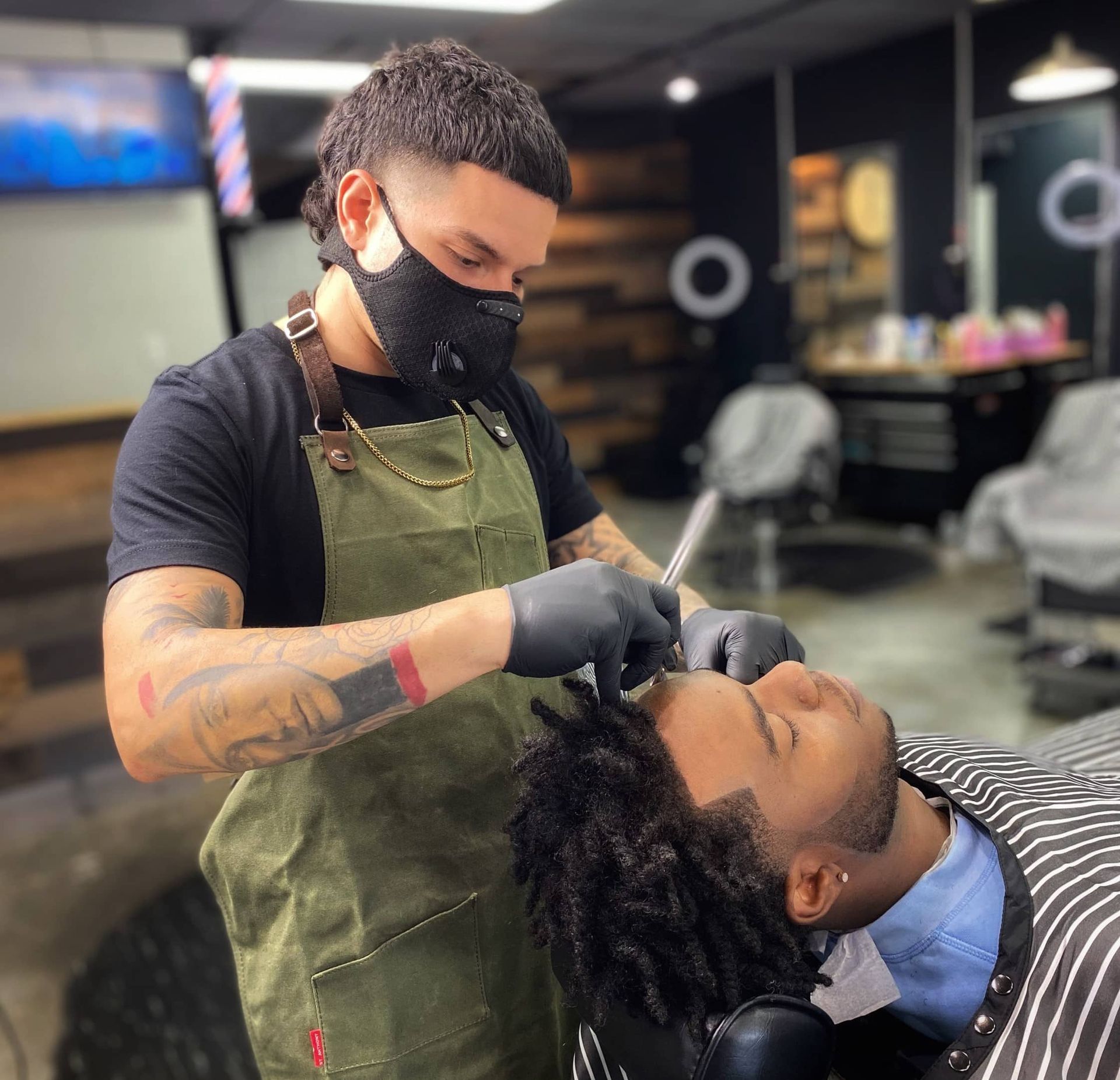 A barber wearing a mask is shaving a man 's face in a barber shop.