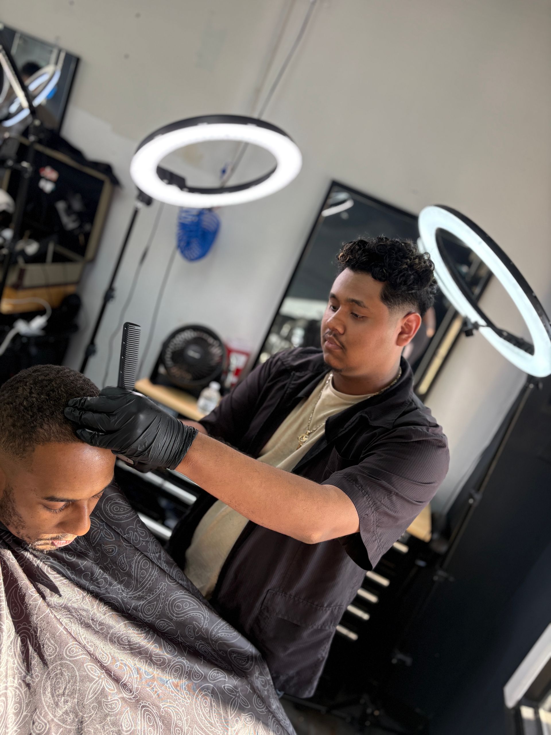 Barber cutting a client's hair in a brightly lit shop. The barber wears black gloves and a black shirt.