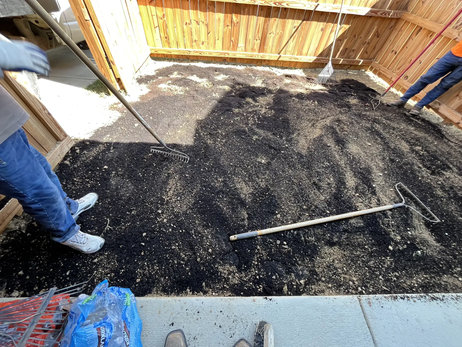 Two workers leveling black gravel in a fenced yard with a long straightedge tool