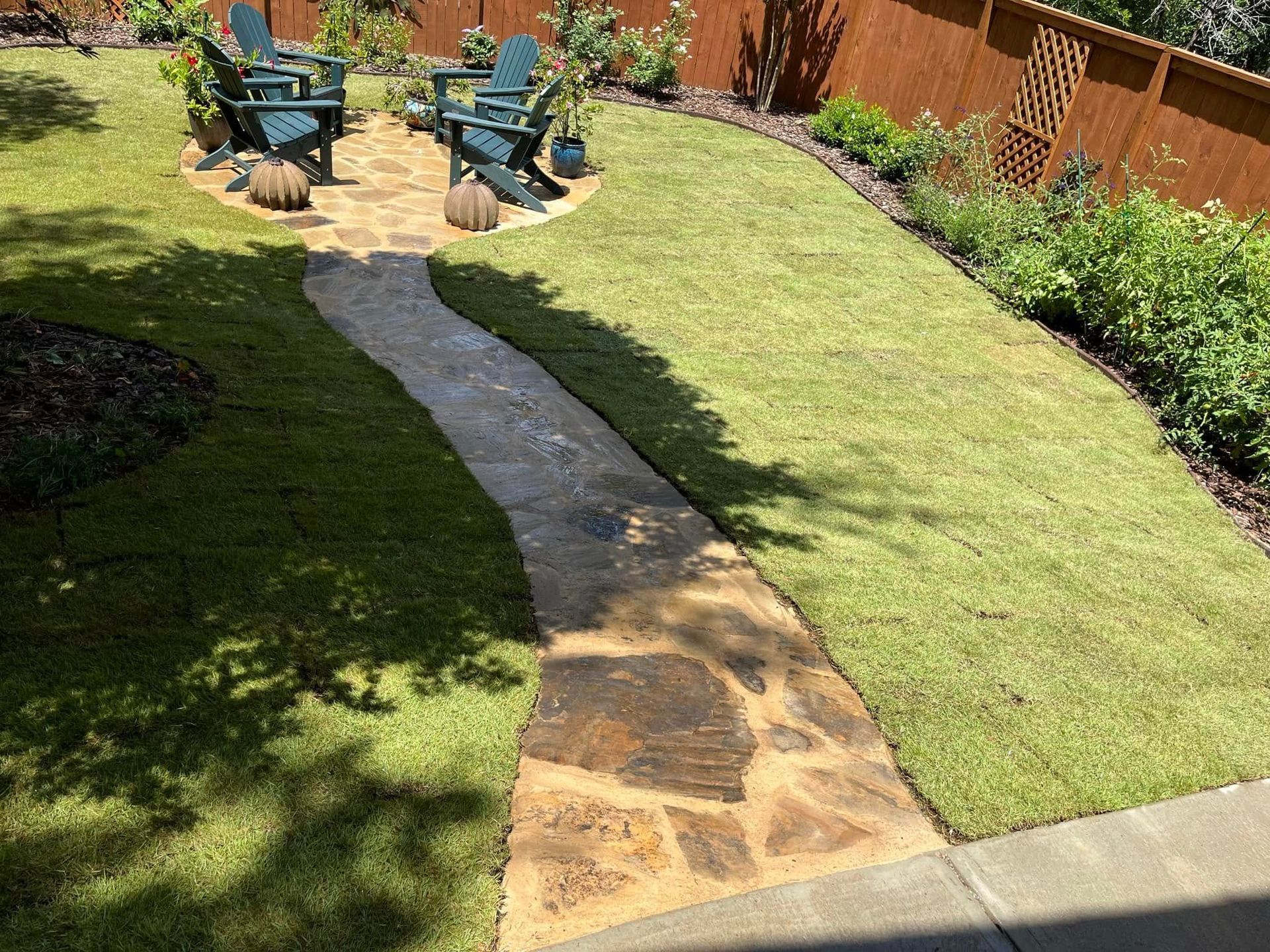 A stone walkway leading to a patio with chairs in a backyard.