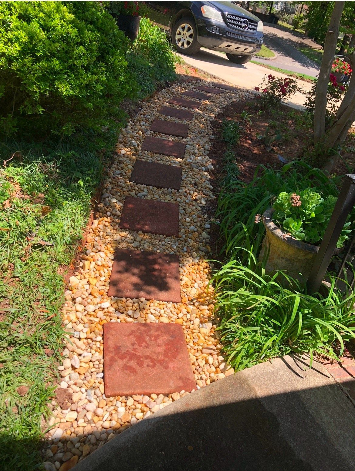 A stone walkway in a garden with a car parked on the side of the road.