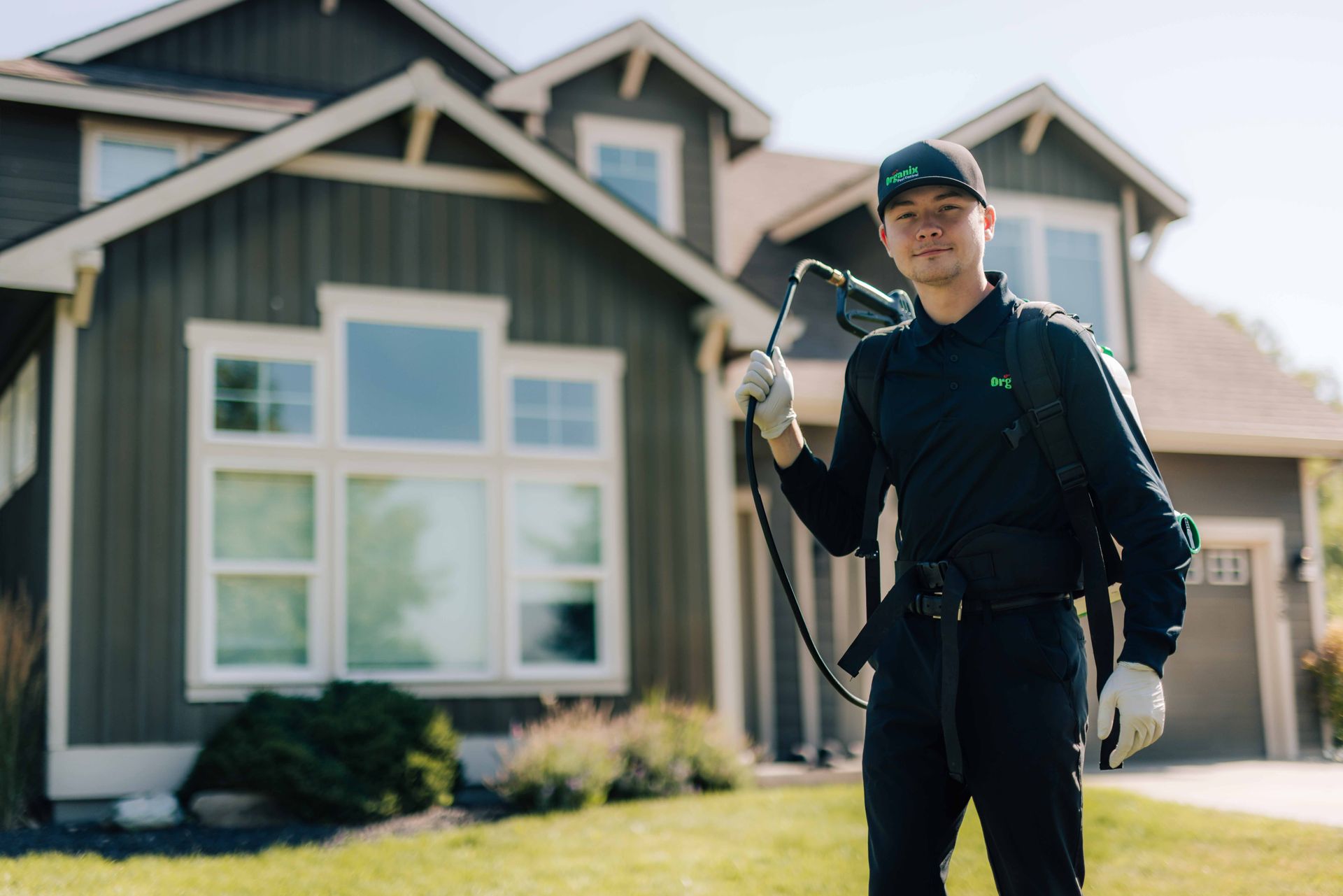 Organix technician spraying a house exterior with a sprayer. He wears a cap, gloves, and a backpack, standing on a lawn.