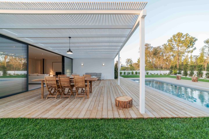 a wooden deck with a table and chairs under a pergola next to a swimming pool