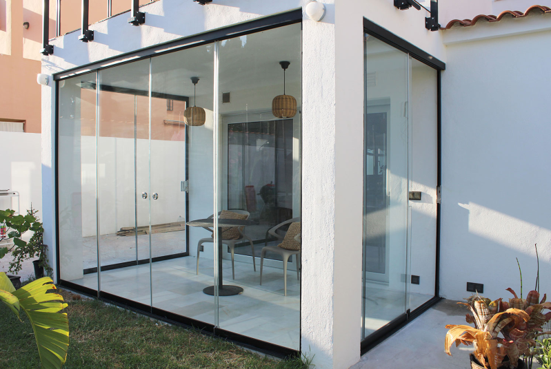 Stanza esterna con vetrate, finiture nere, pareti bianche e lampade in rattan, arredata con tavolo e sedie.