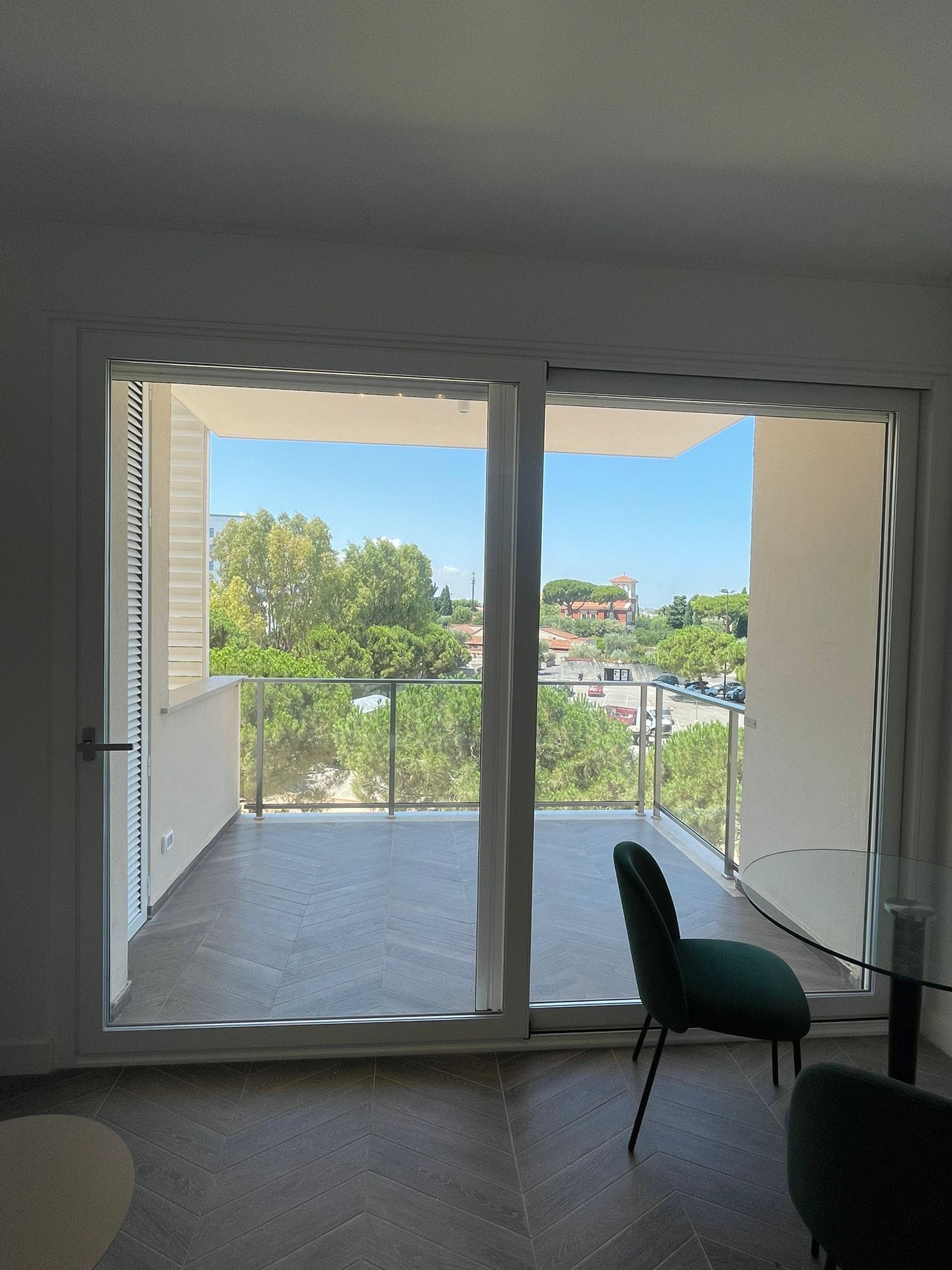 Camera luminosa con porta scorrevole in vetro che si apre su un balcone con alberi verdi e vista sul cielo azzurro. All'interno è presente una sedia color verde acqua.