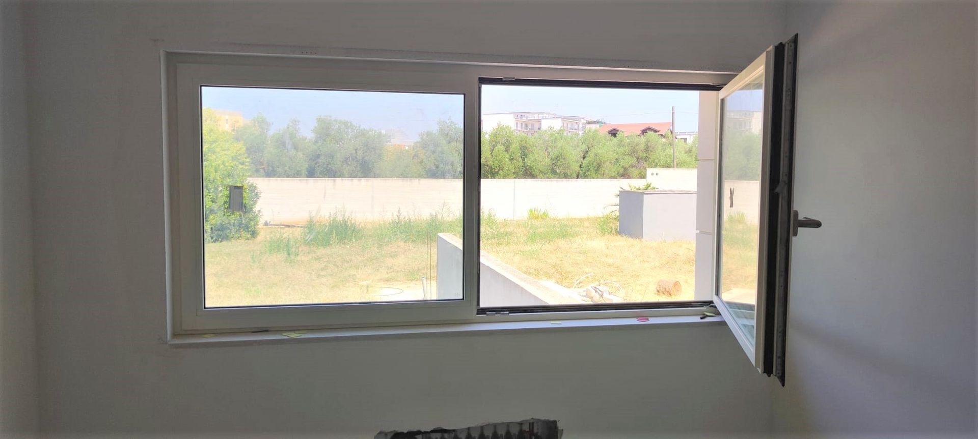 Finestra aperta in una stanza bianca, con vista sull'erba verde, sugli alberi e su un edificio in lontananza.