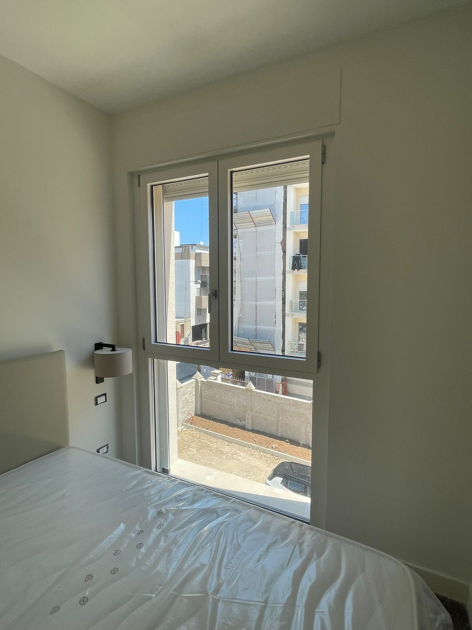 Camera da letto con finestra che si affaccia su un'area esterna e su un edificio; in primo piano c'è un letto.