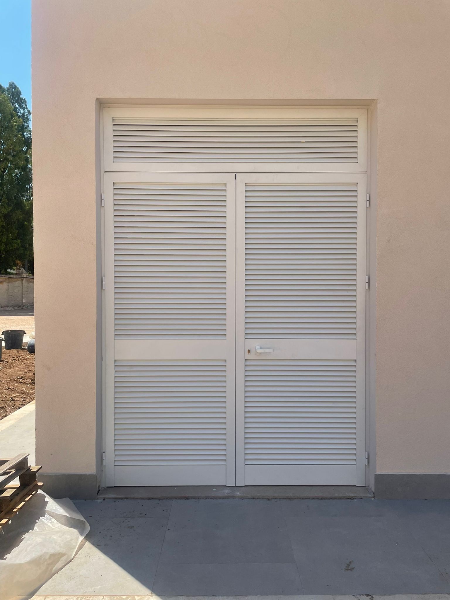 Porte doppie bianche con persiana e finestra a traverso abbinata. Esterno dell'edificio, pavimento in cemento.