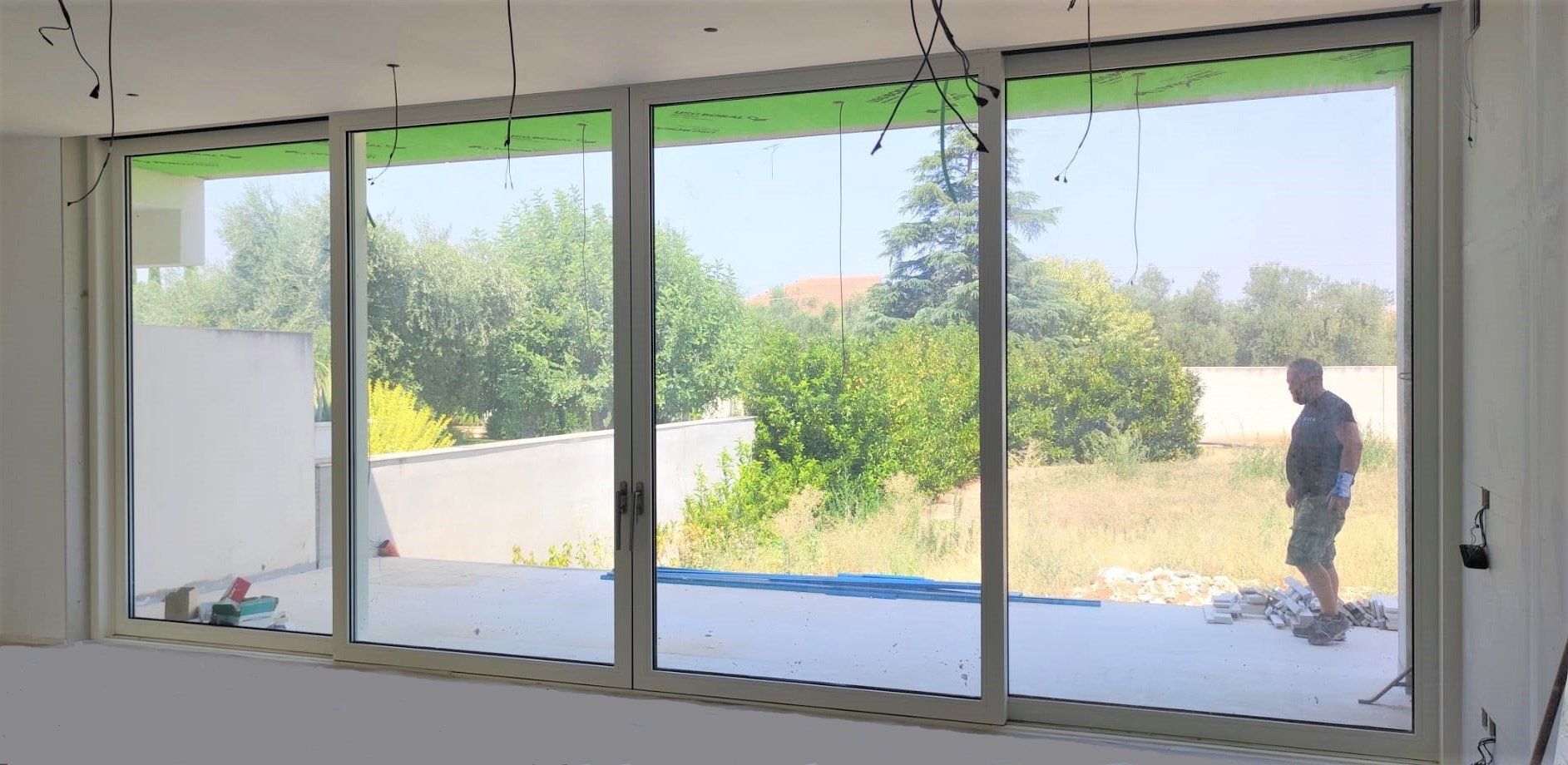 Un uomo in piedi fuori da una grande porta scorrevole in vetro, osserva un cortile in una giornata di sole. Interno di una casa incompiuta.