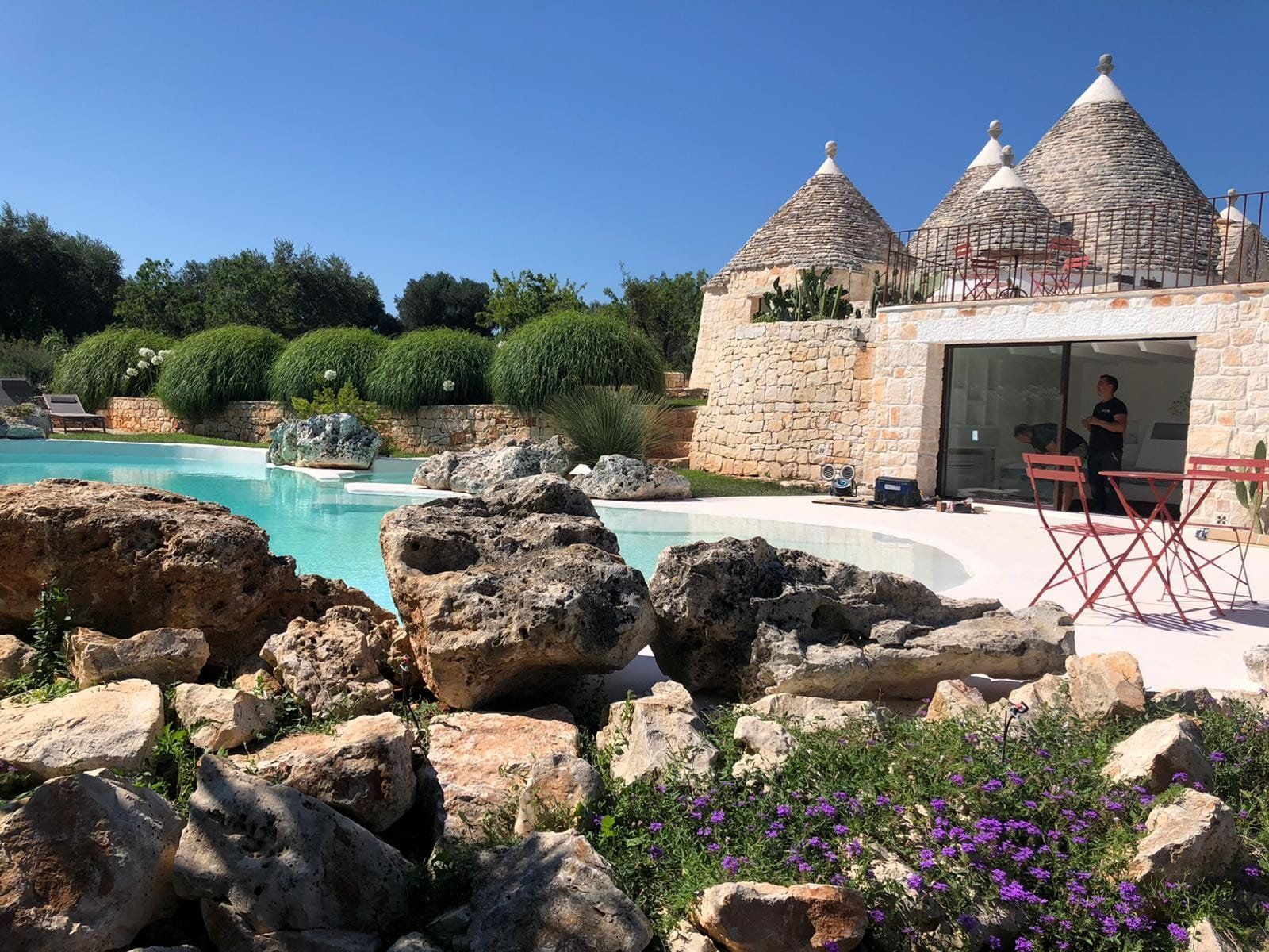 Trulli con tetti conici accanto a una piscina con grandi rocce e fiori viola. Giornata di sole.