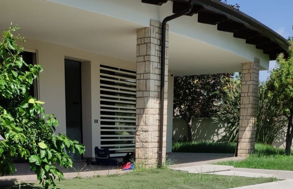 Portico con colonne in pietra beige di una casa con pareti bianche, circondato da alberi e da un sentiero lastricato.