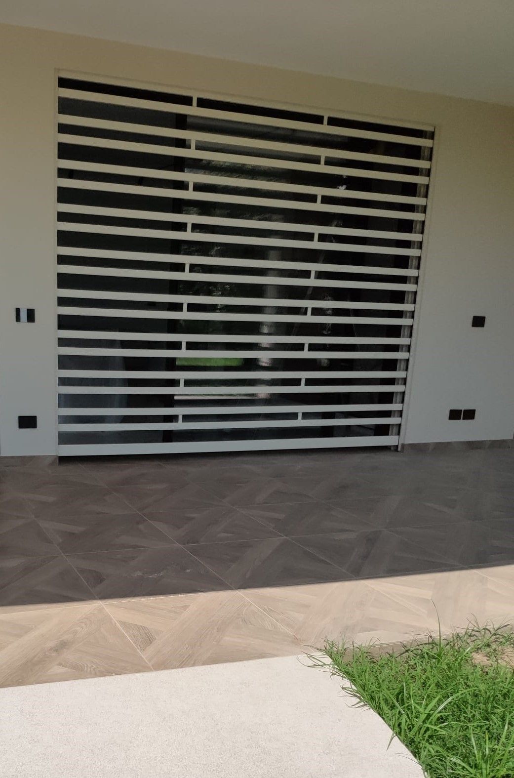 Persiana di sicurezza a stecche bianche che copre una finestra, su una parete beige, con pavimento piastrellato ed erba.