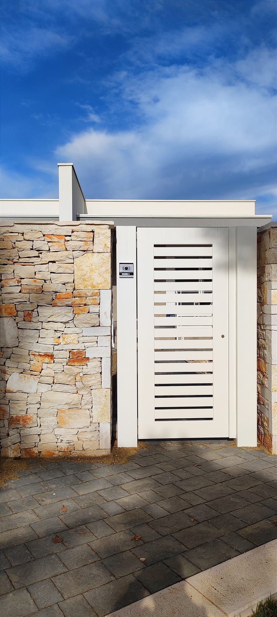 Cancello bianco con stecche orizzontali, muro di pietra, cielo azzurro.