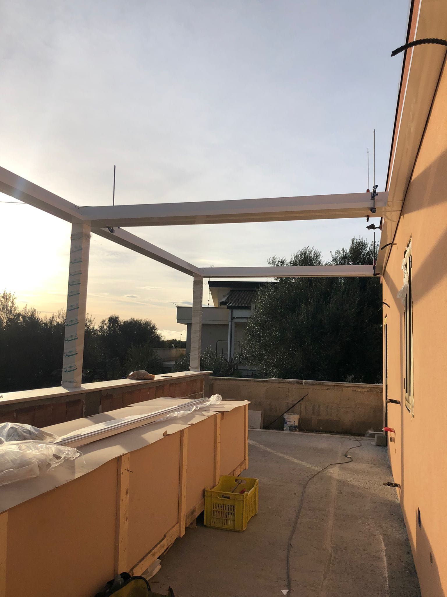 Un pergolato bianco parzialmente costruito su un tetto con vista su alberi ed edifici, con un muro beige sulla destra.