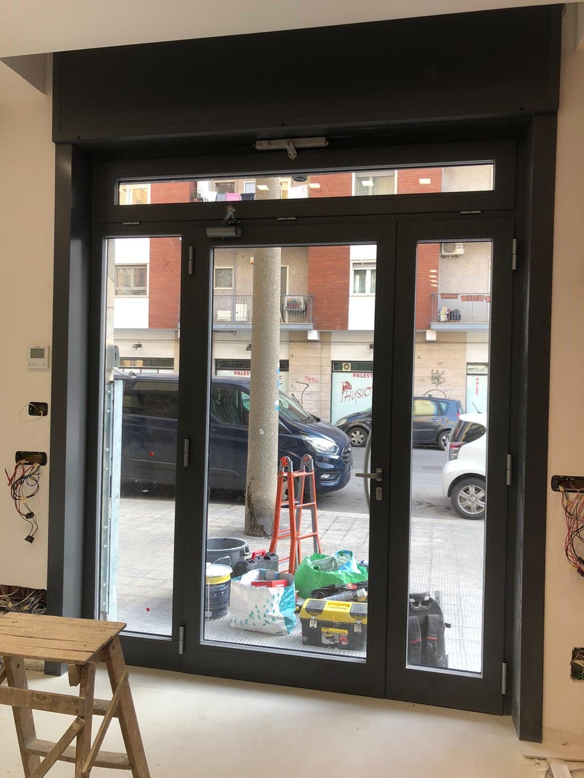 Porta a vetri con cornice scura, aperta su una strada cittadina con auto parcheggiate e un albero. Una scala è in primo piano.