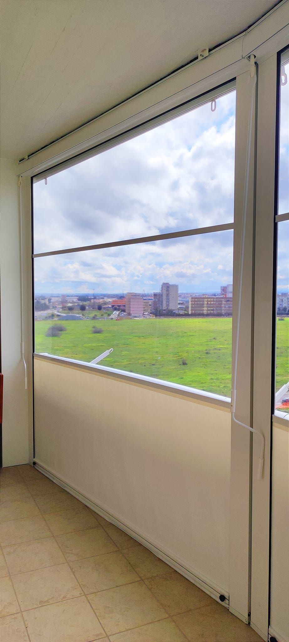 Vista da una finestra che si affaccia su un campo verde e su edifici sotto un cielo nuvoloso.