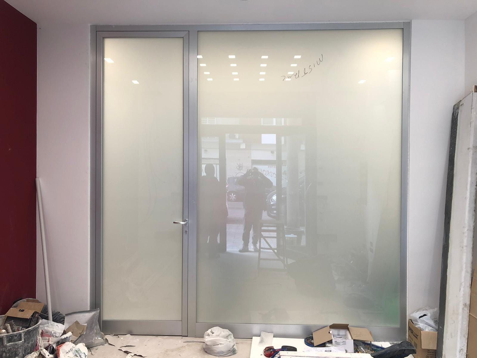 Porta e pannello laterale in vetro smerigliato. Due figure visibili all'interno. Detriti di costruzione sul pavimento.