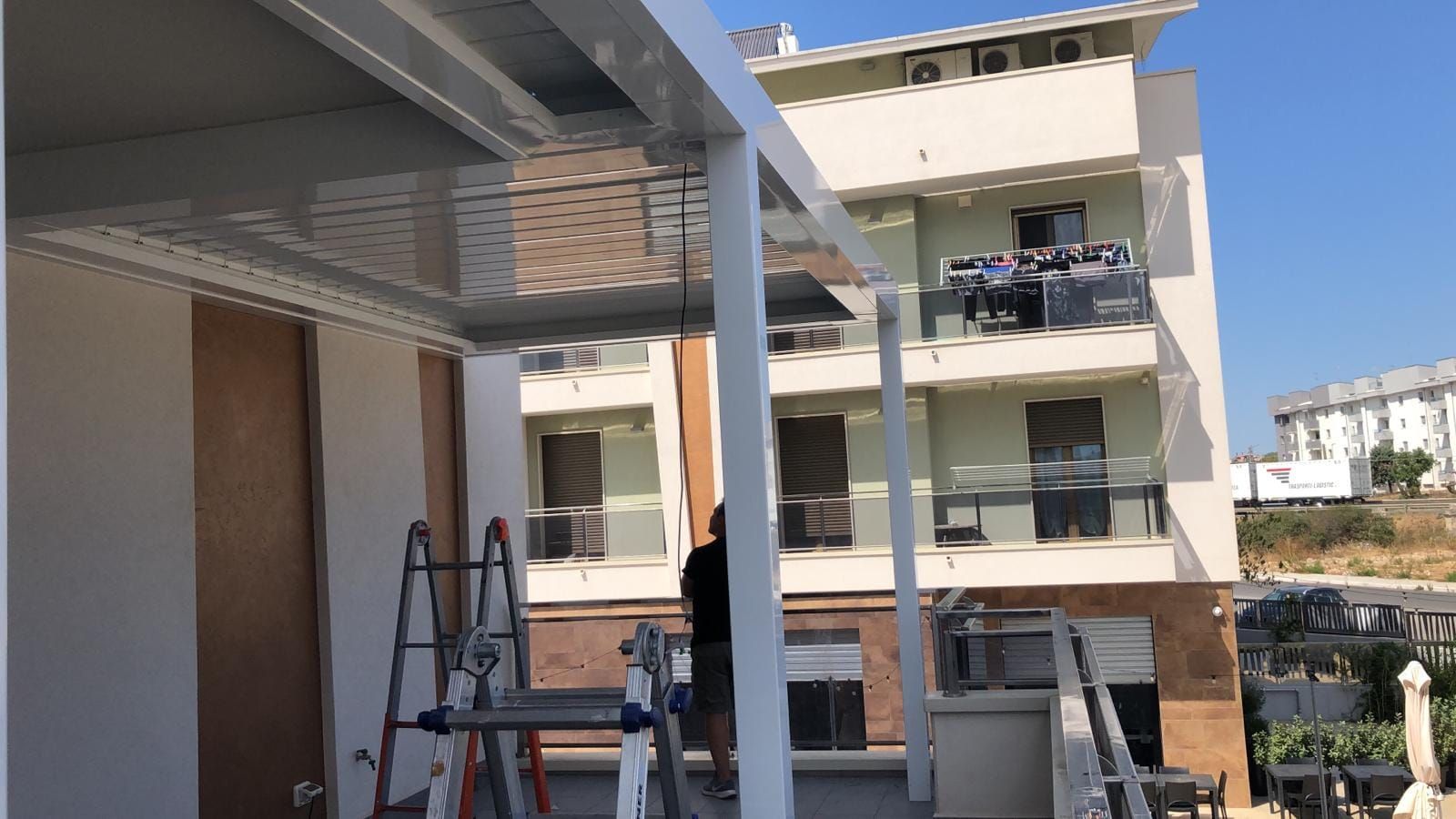 Uomo su una scala che installa una tenda da sole in metallo bianco sul balcone di un edificio; giornata di sole.
