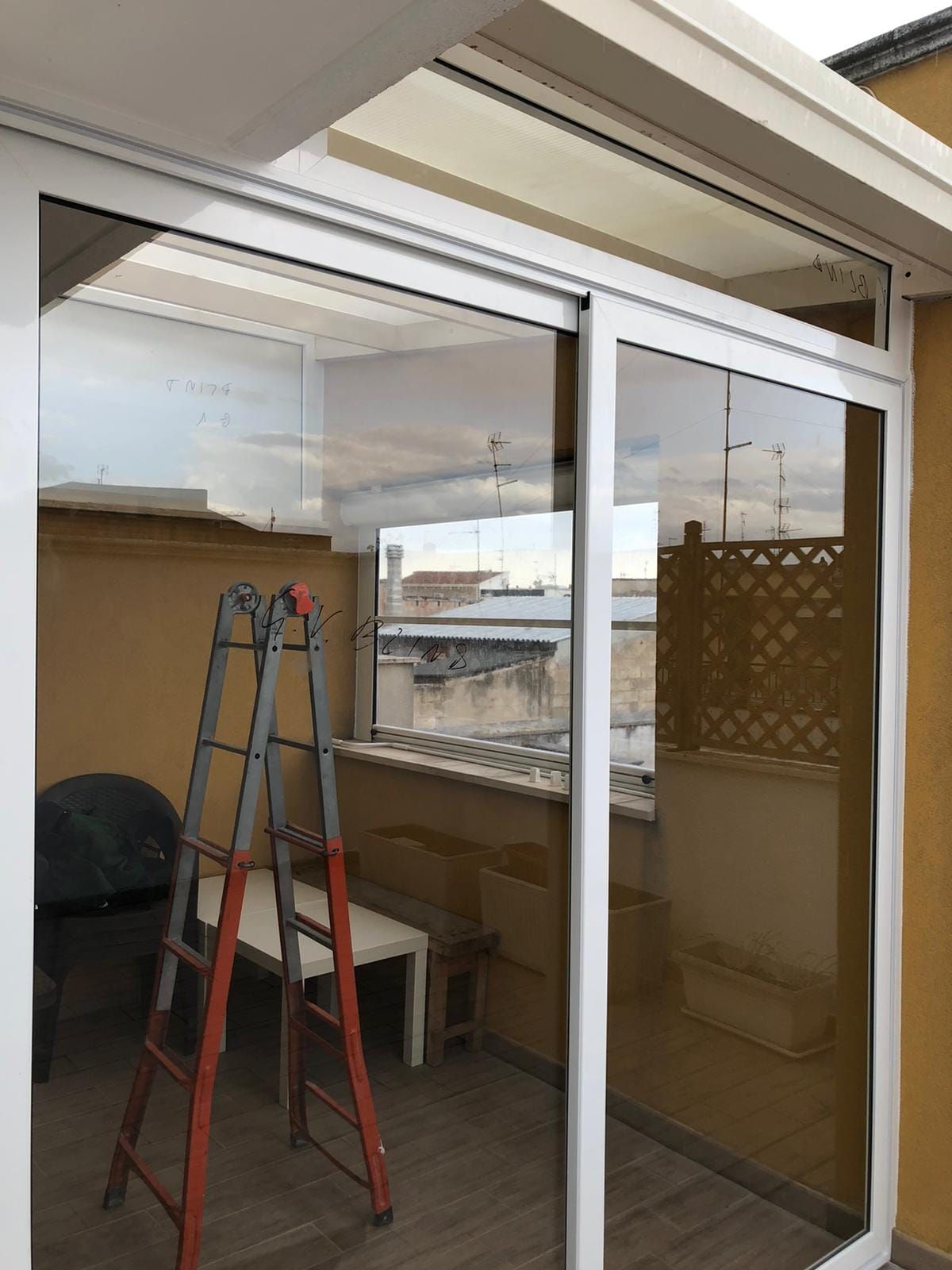 Porte scorrevoli in vetro su un balcone con una scala e un tavolino all'interno. Cielo coperto riflesso.