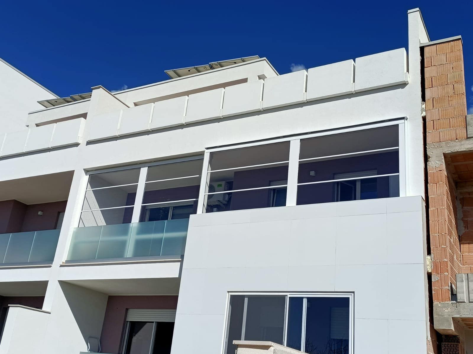 Edificio bianco con finestre in vetro e balconi contro il cielo azzurro; mattoni incompiuti visibili sulla destra.