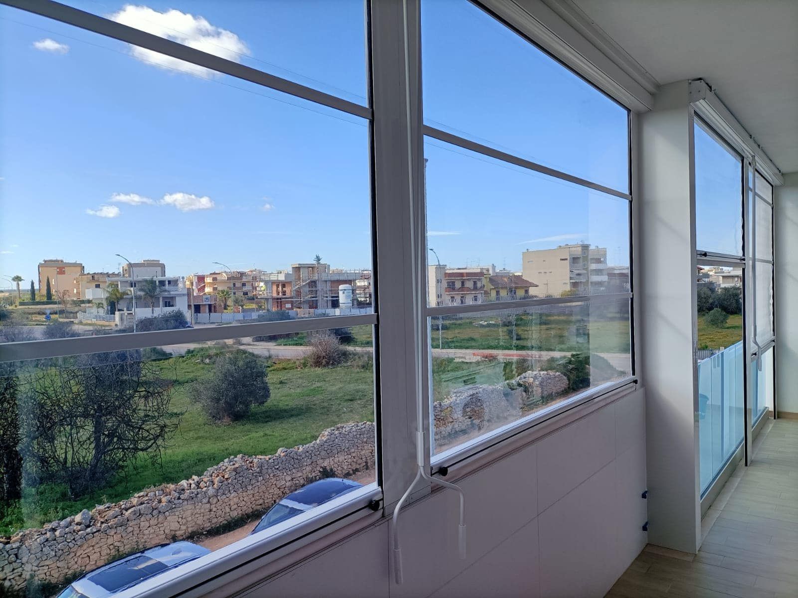 Vista dal balcone chiuso che domina una città con cielo soleggiato e paesaggio verde.
