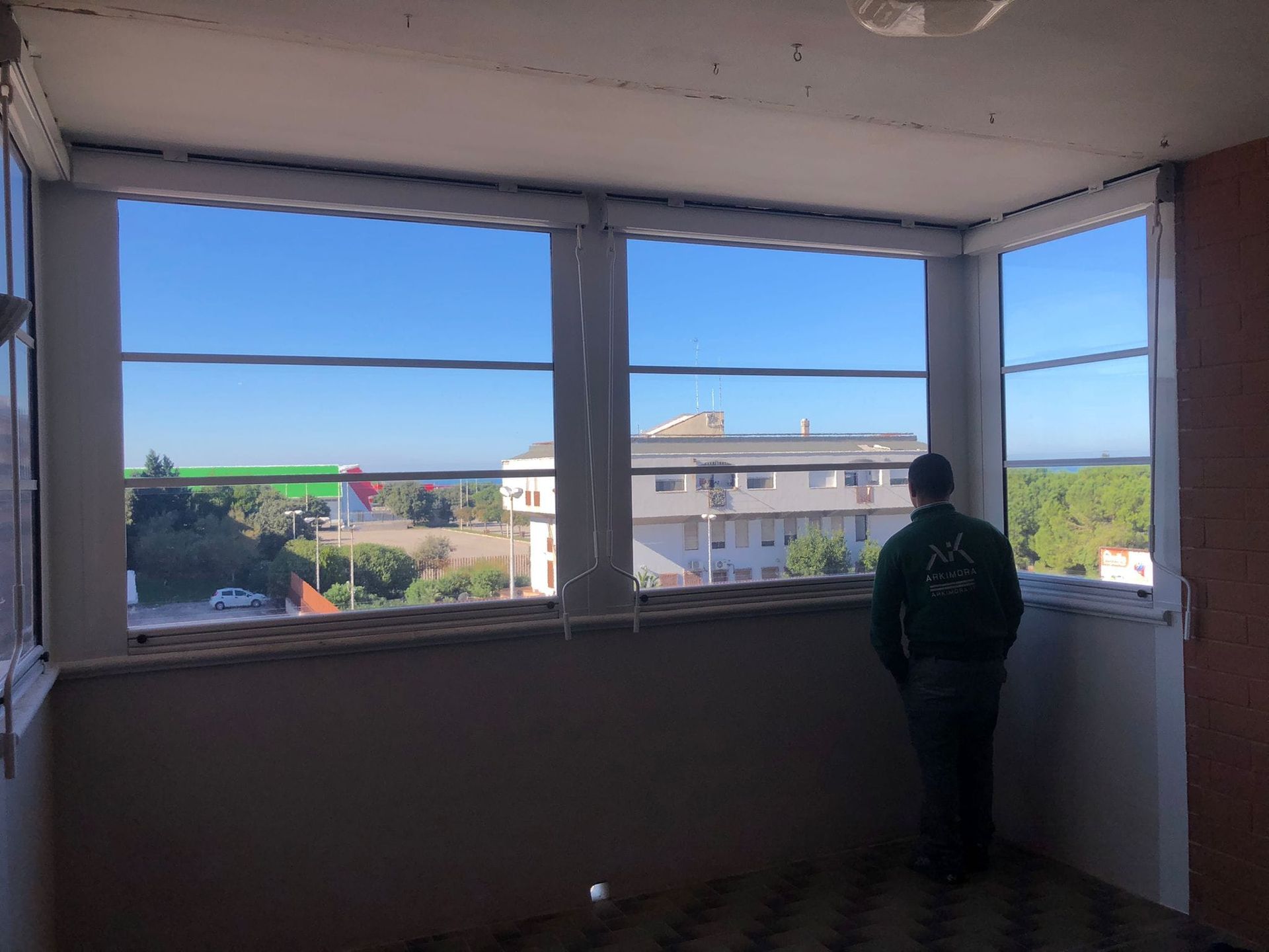 Persona che guarda fuori dalla finestra di una veranda verso una città con cielo azzurro.