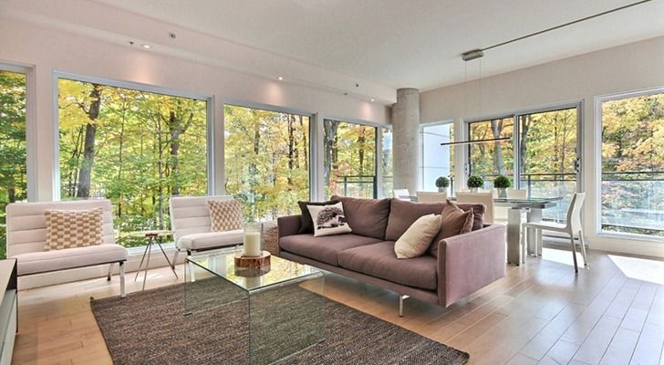 Soggiorno con ampie finestre con vista sugli alberi. Divano marrone, sedie bianche, tavolino in vetro e pavimento in legno.