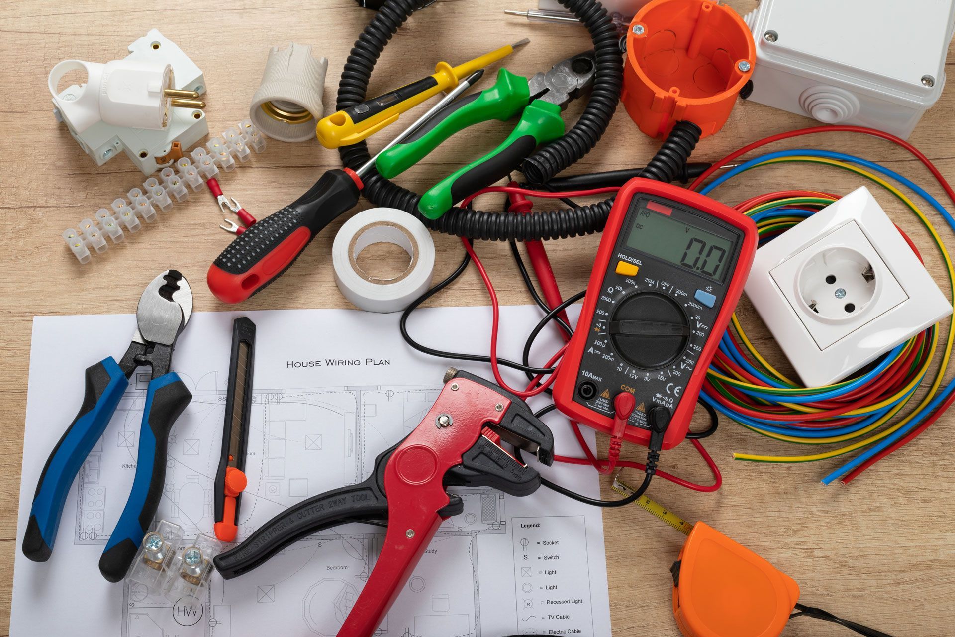Assorted electrical tools and supplies on a workbench, including pliers, wire, tape, outlet, and multimeter.