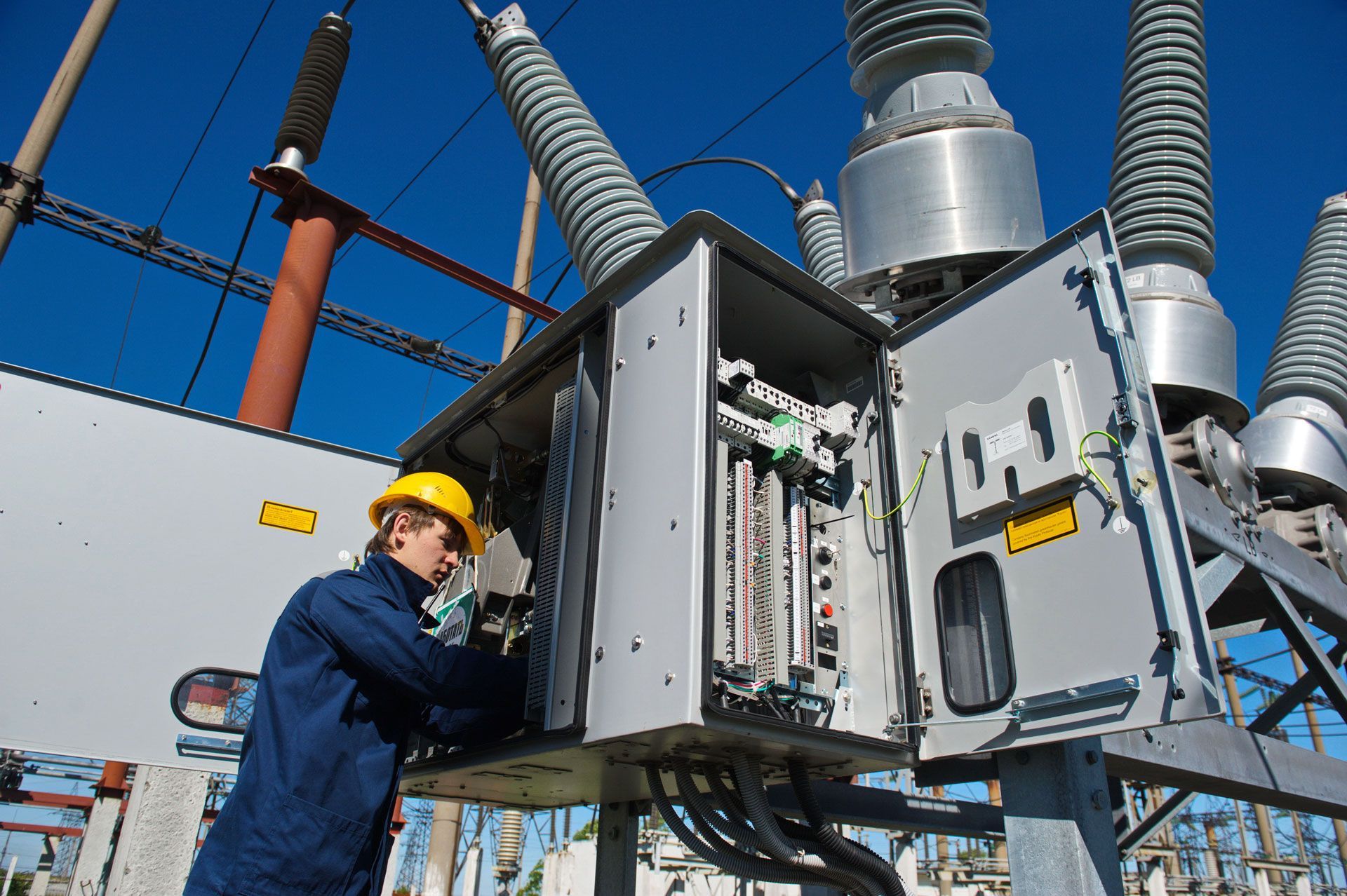 Technician in hard hat working on an open electrical control panel at a substation