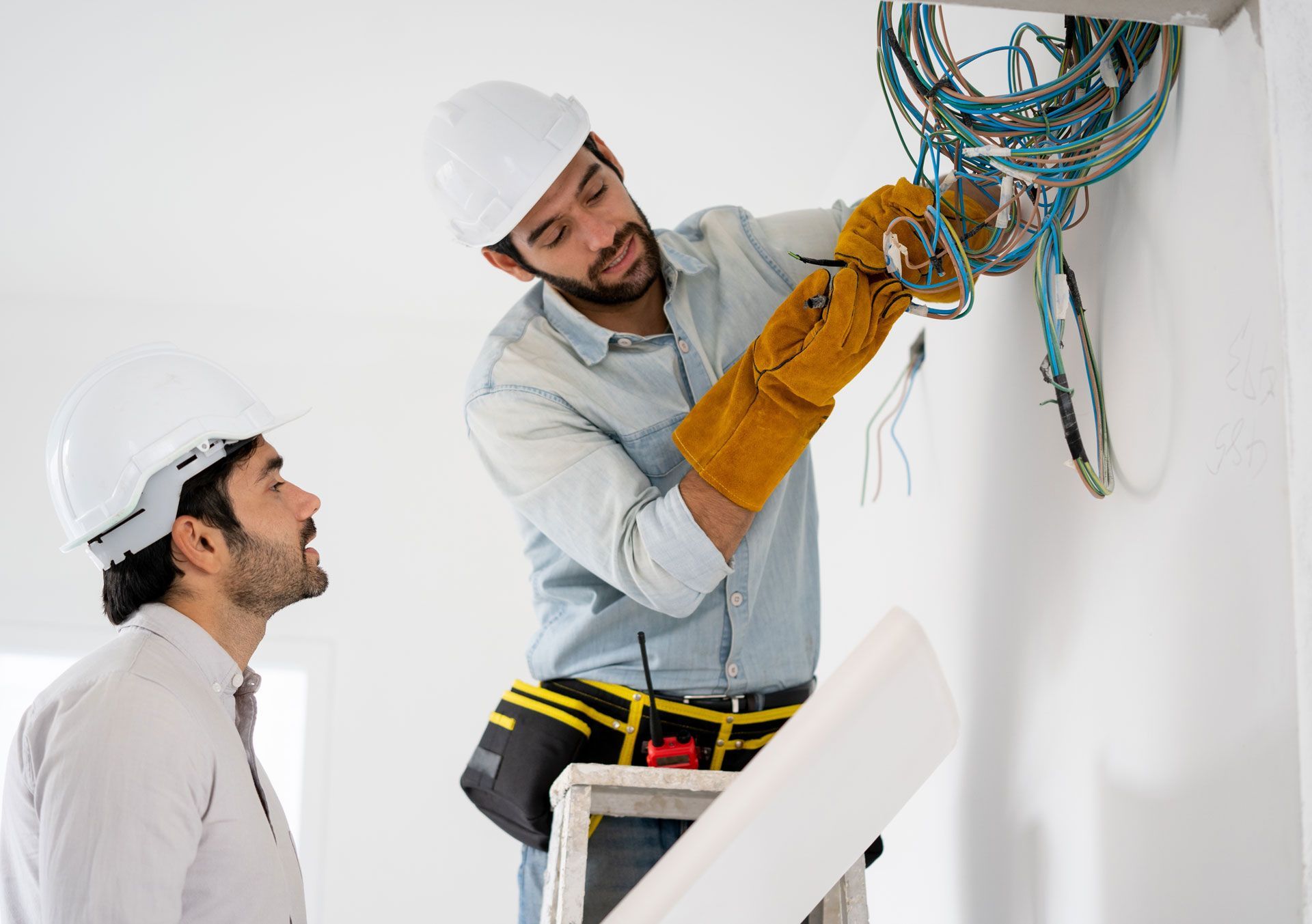 Two electricians inspecting exposed wiring on a wall, one standing on a ladder.