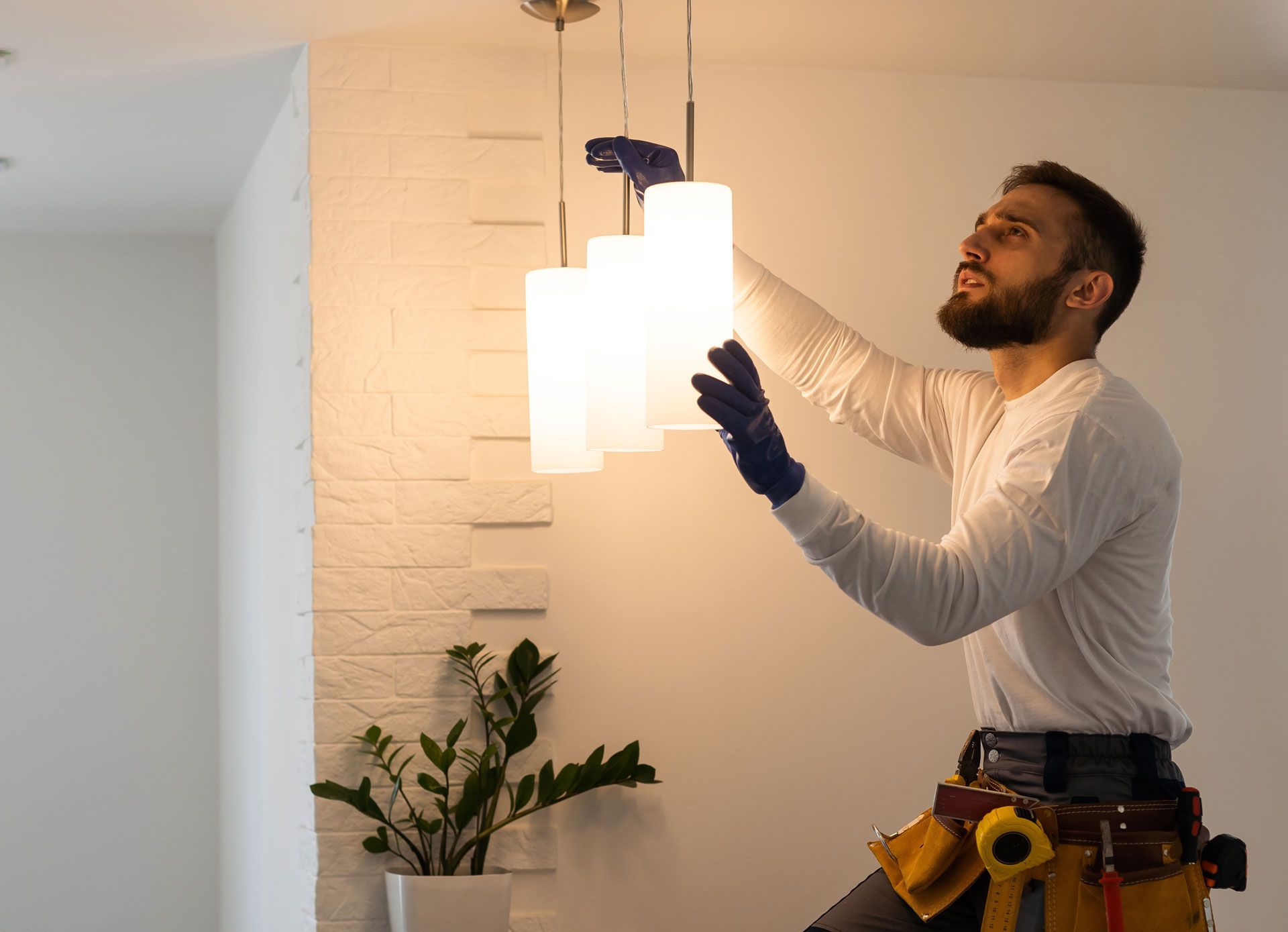 Man installing a white pendant light fixture indoors, reaching up beside a brick wall and plant.
