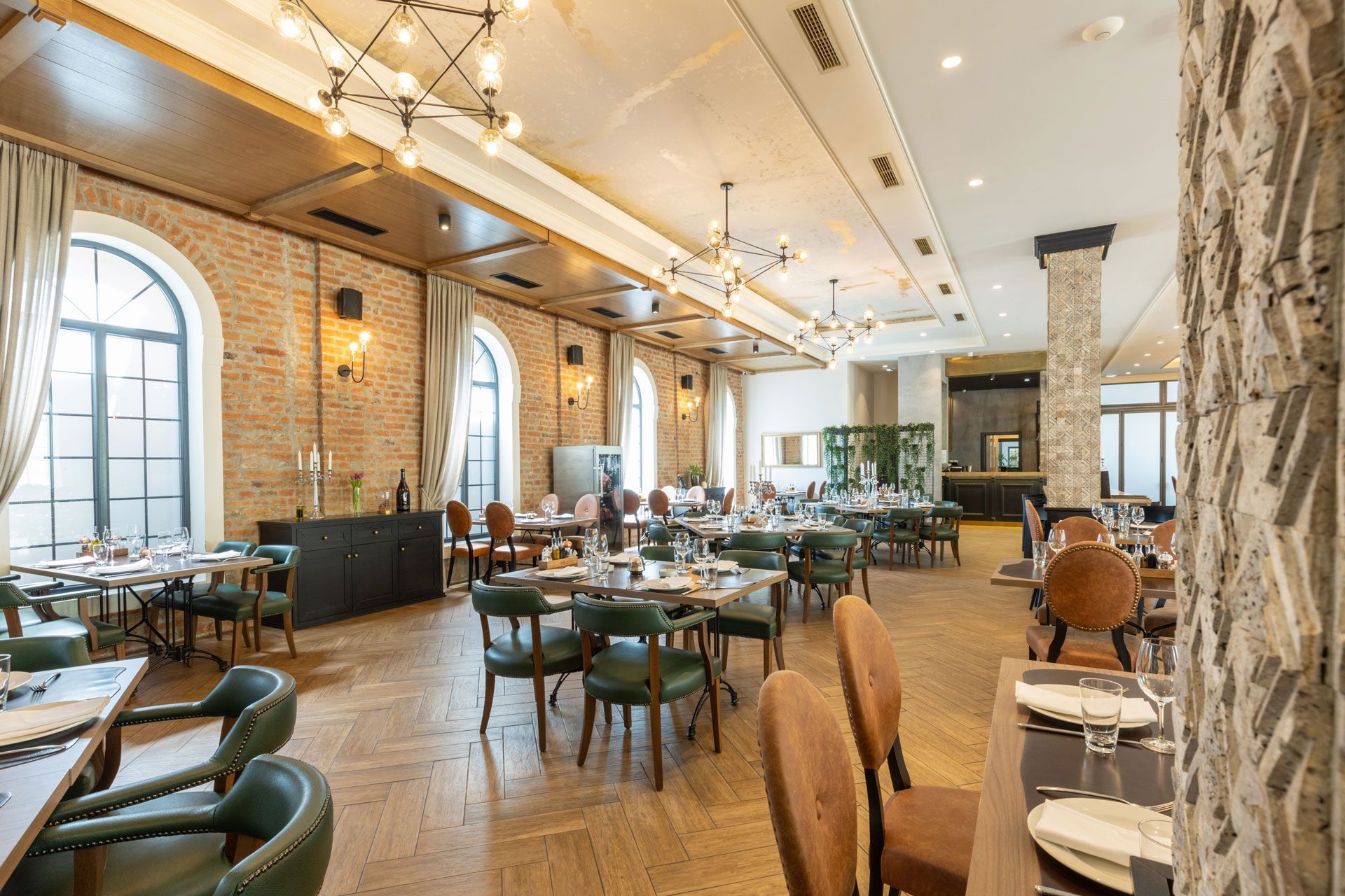 Bright, modern restaurant interior with wooden tables, green chairs, brick walls, and high arched windows.