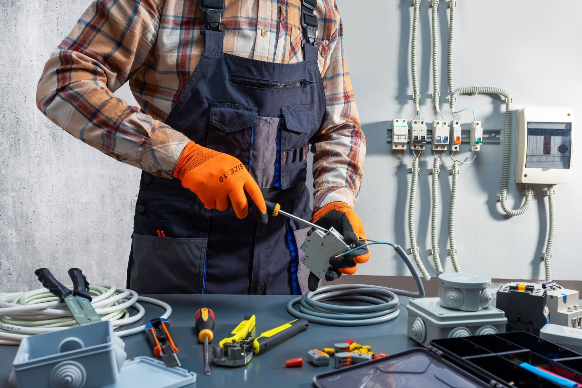 Electrician in orange gloves wiring cables at a workbench with tools and wall-mounted electrical panels
