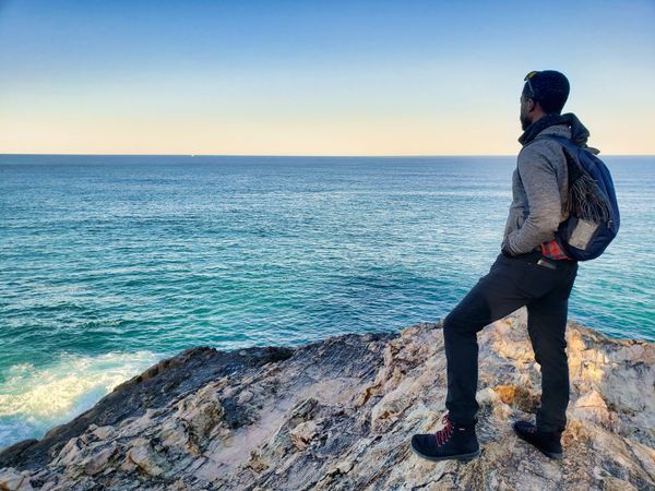 A young Black person standing atop a cliff looking out towards the ocean.