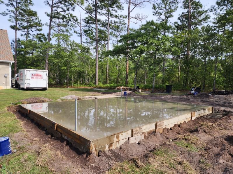 Freshly poured concrete slab in a wooded area, surrounded by wooden forms and a service truck.