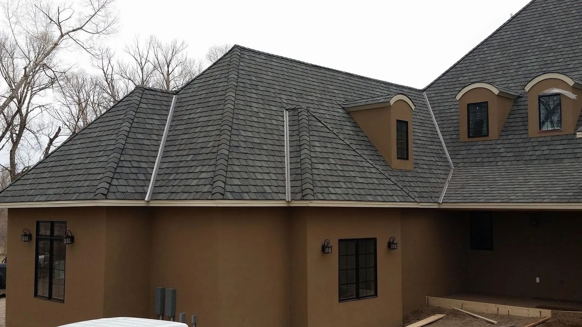 Brown stucco house with a dark gray shingle roof, windows, and a snow-covered area.