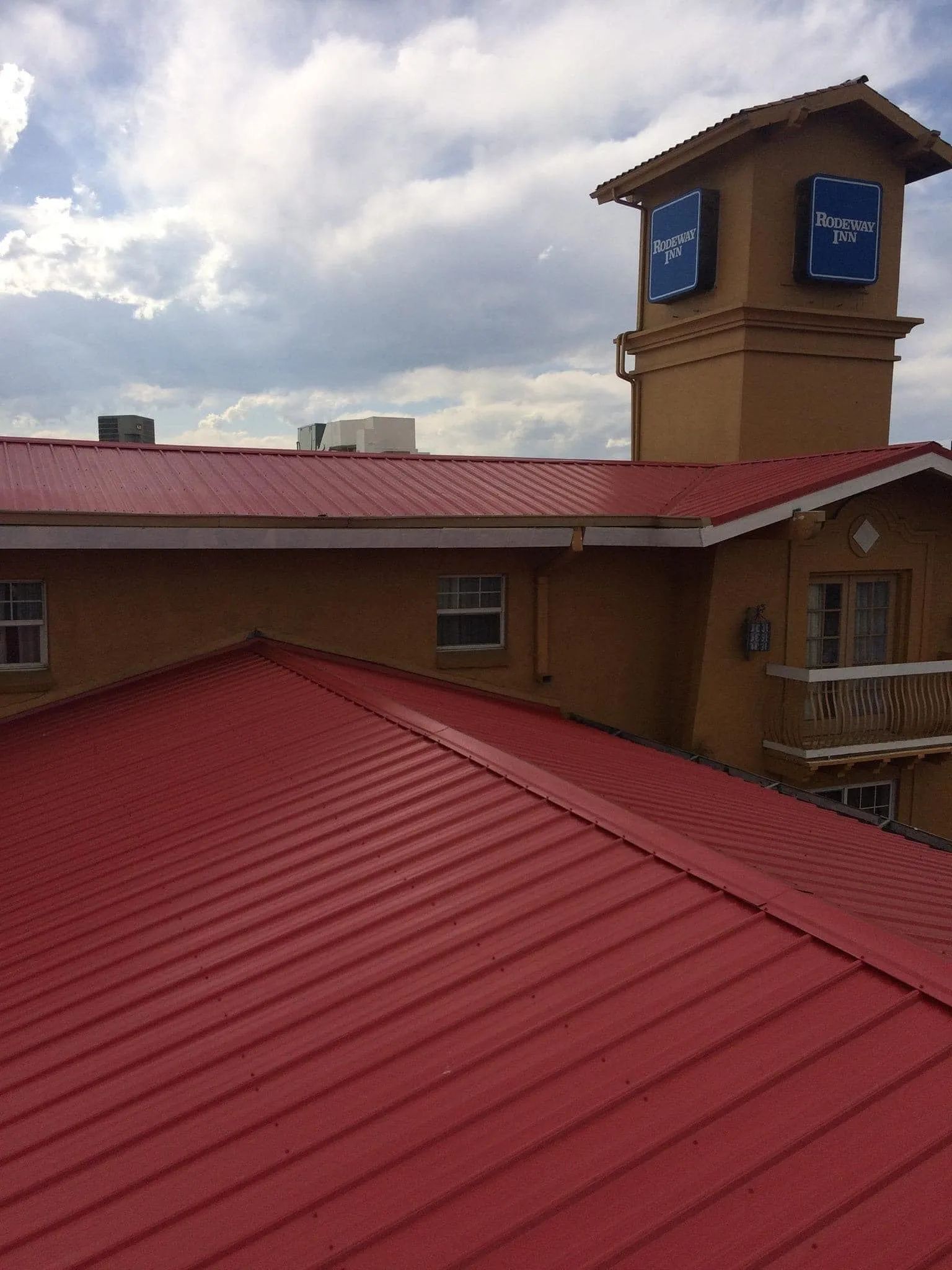 Red corrugated metal roof on a tan building with a tower, blue signs, and a cloudy sky.